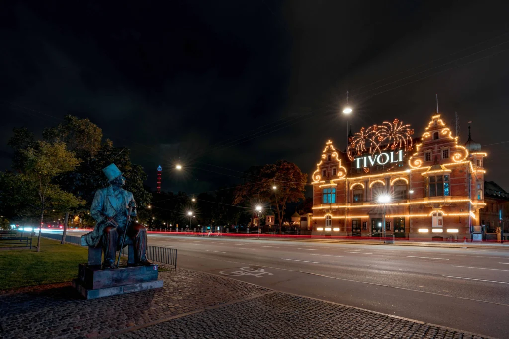 The iconic entrance to Tivoli Gardens in Copenhagen, lit up with festive lights and decorations, featuring a statue in the foreground.