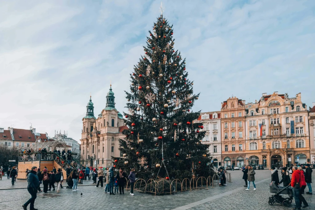 A towering Christmas tree adorned with red and gold ornaments in Prague's Old Town Square, surrounded by festive market stalls.