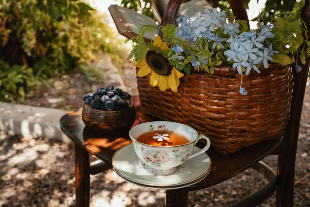 A beautifully arranged tea setup on a wooden table featuring a floral teacup, fresh blueberries in a bowl, and a basket of flowers, set in a tranquil outdoor garden