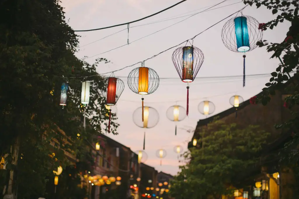 Colorful lanterns hanging above a street at dusk, with warm lights glowing and a peaceful atmosphere set against a backdrop of trees and buildings.