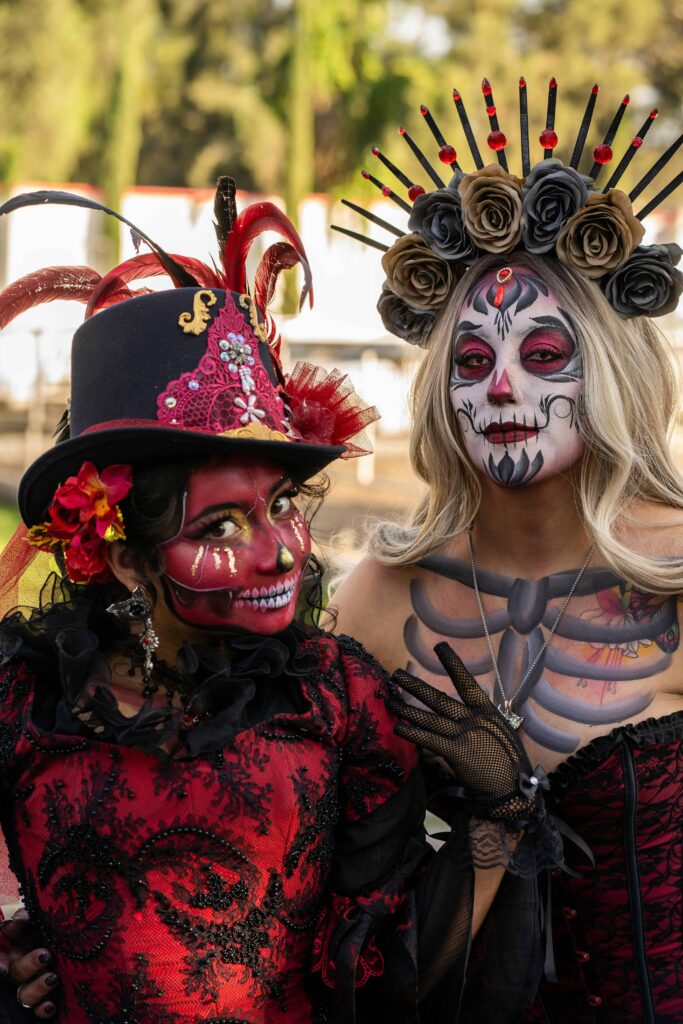 Two women dressed in elaborate Day of the Dead costumes, with intricate face paint and vibrant accessories, posing together at a cultural festival.