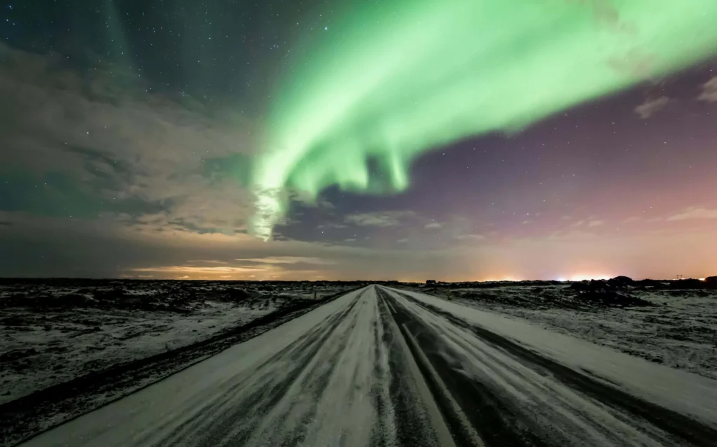 Long Icelandic road under the Aurora lights, with snowy terrain and a glowing sky.