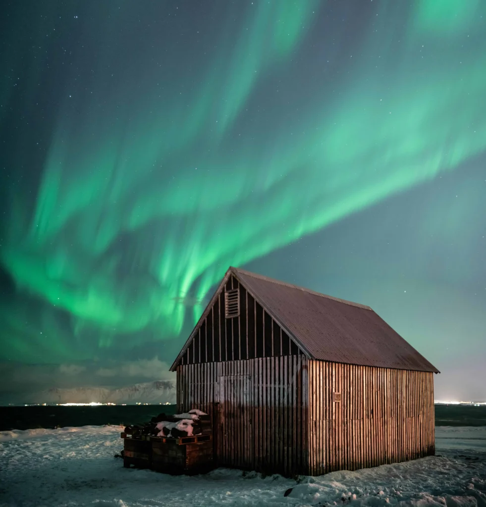 Northern lights illuminating a rustic wooden cabin on a snowy Icelandic plain.