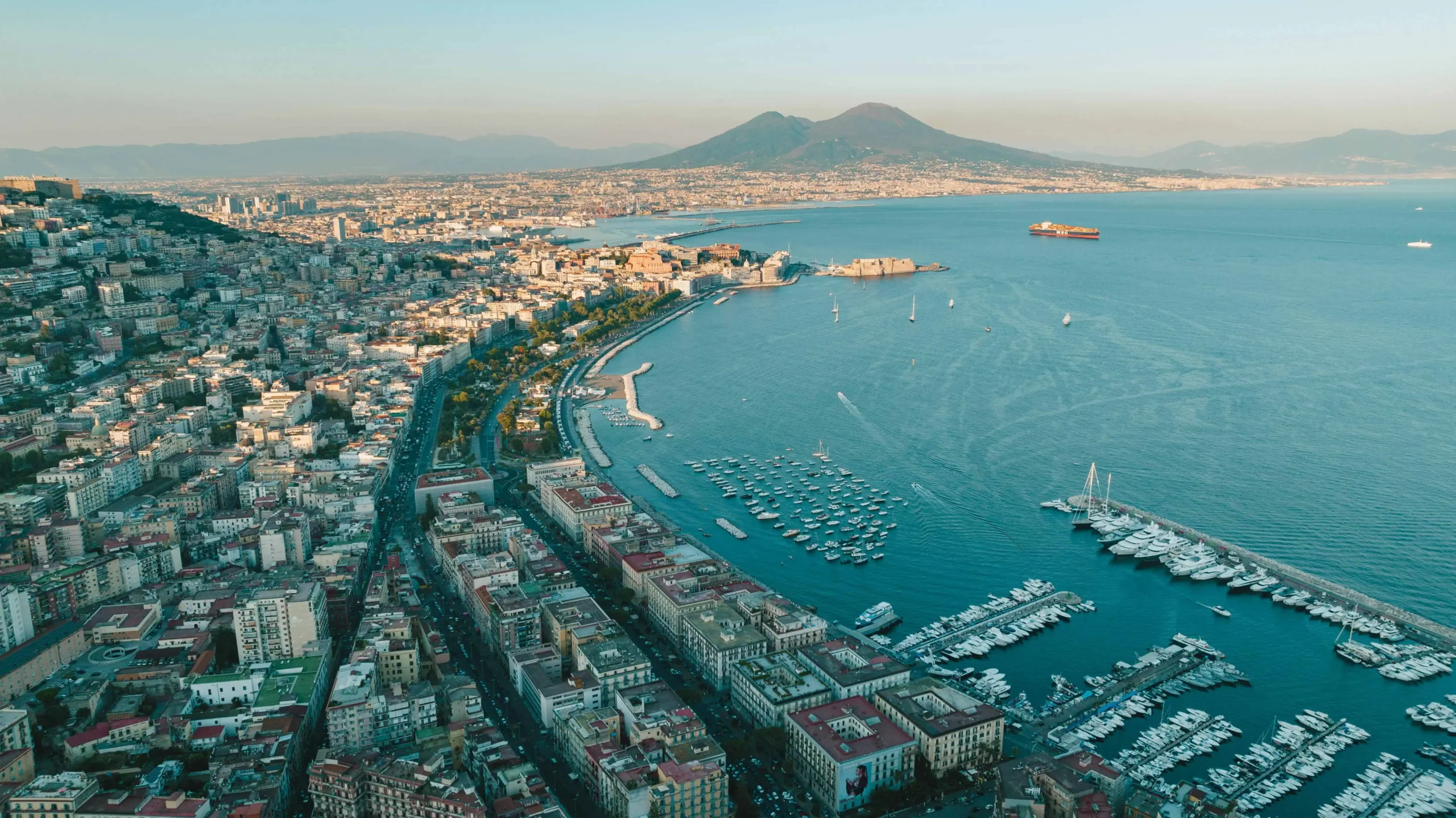 An aerial view of Naples, Italy, featuring the bustling cityscape, the Bay of Naples, and the iconic Mount Vesuvius in the background, with yachts docked along the coastline