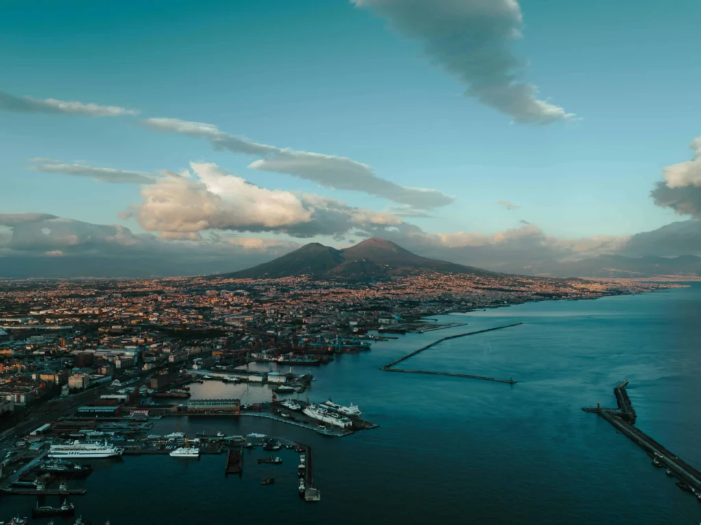 A stunning aerial view of Naples, Italy, featuring the iconic Mount Vesuvius in the background, with the vibrant cityscape and coastline under a serene blue sky.