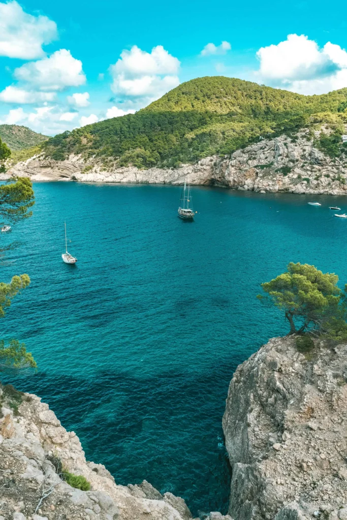 Stunning cove in Spain with turquoise waters, sailboats, and lush green hills, offering a peaceful escape for visitors.