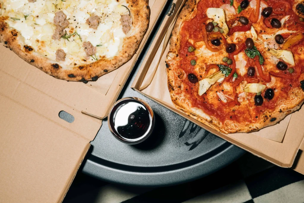 Two authentic Neapolitan pizzas served in cardboard boxes, one topped with creamy mozzarella and sausage, and the other with tomato, olives, and artichokes, paired with a glass of red wine