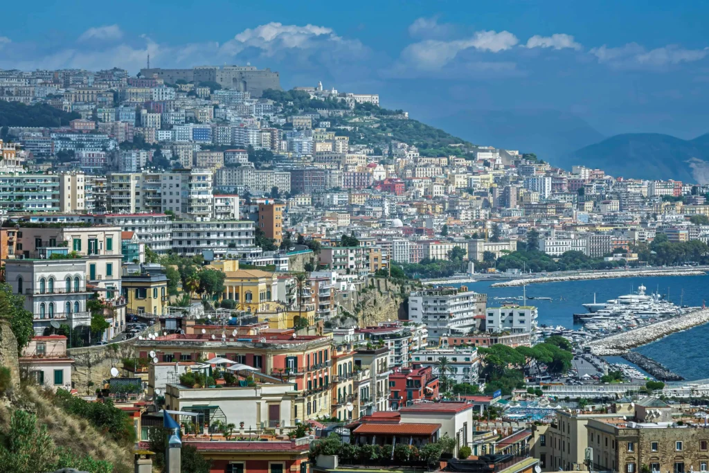 A vibrant view of Naples, Italy, featuring colorful buildings along the coastline with the sparkling blue sea and marina filled with yachts in the background.