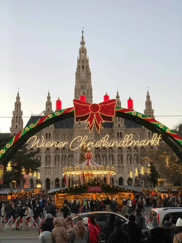 The illuminated entrance to the Vienna City Hall Christmas Market, featuring an arch with red bows, a carousel, and vibrant holiday stalls.