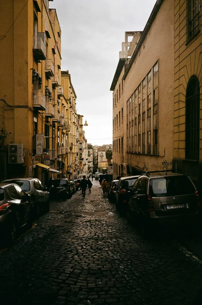 A charming cobblestone street in Naples, Italy, flanked by colorful historic buildings and parked cars