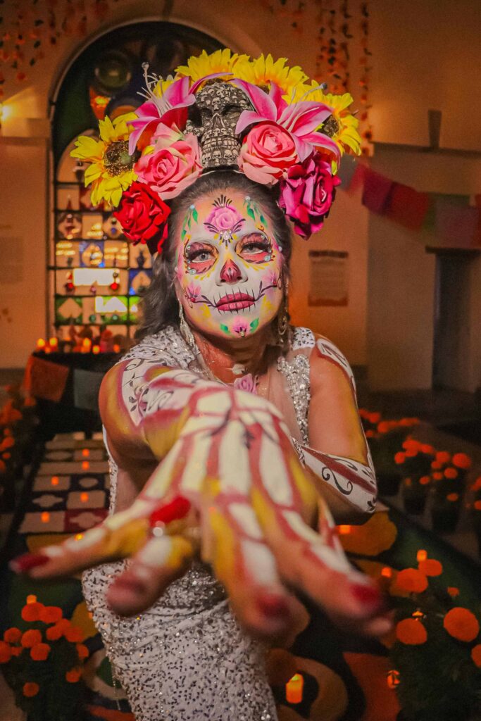 A woman dressed in traditional Day of the Dead makeup and attire, with colorful flowers and vivid face paint, extending her hand toward the camera in Huatulco Mexico