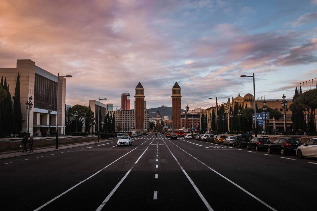 Wide avenue leading to the Venetian Towers at Plaça d'Espanya in Barcelona, captured under a dramatic sky during the golden hour, with a view towards Montjuïc Hill in the distance.