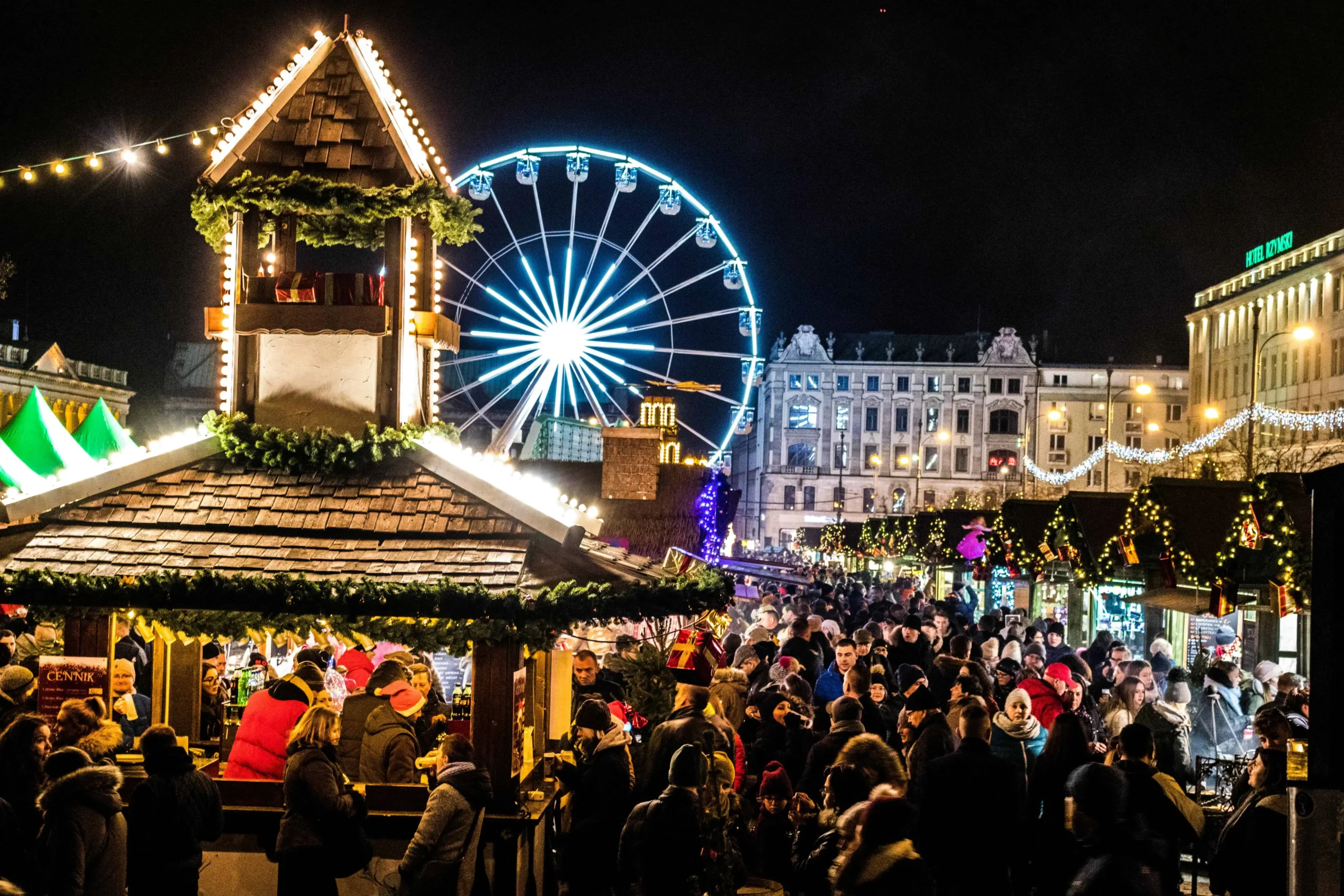 A vibrant European Christmas market at night, featuring a brightly lit Ferris wheel in the background, festive stalls decorated with greenery and lights, and a lively crowd enjoying the holiday atmosphere. Perfect example of the best Christmas markets in Europe.