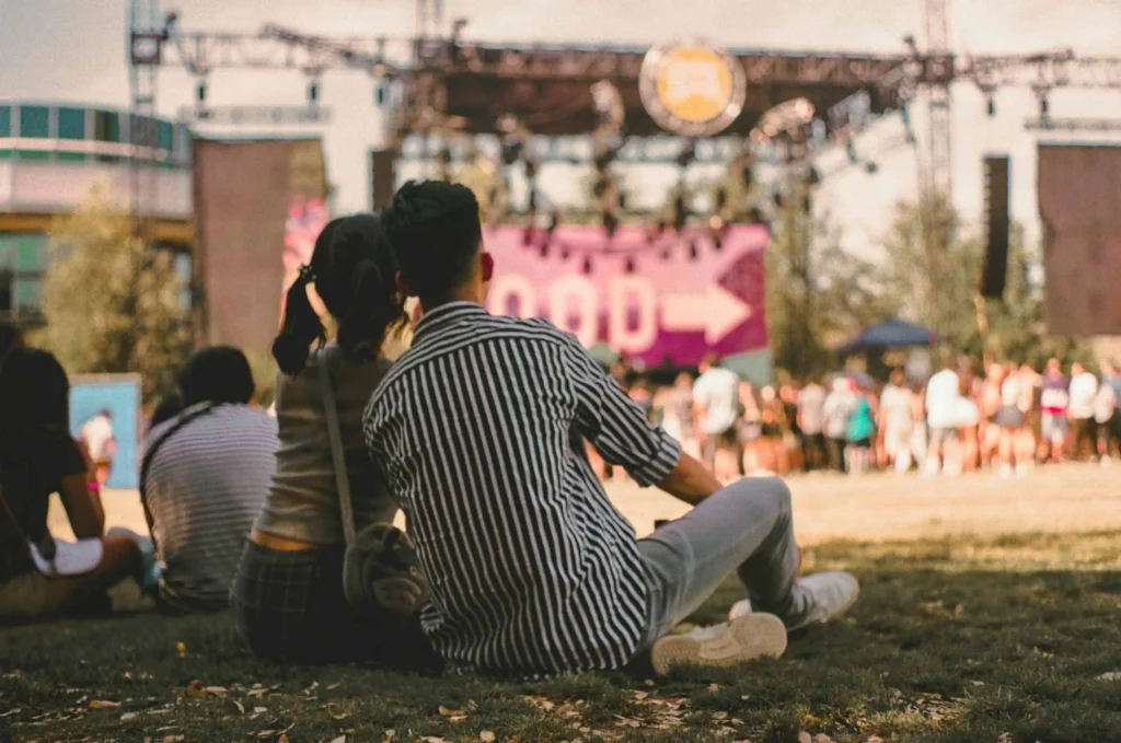 A couple sitting on the grass, enjoying a live jazz performance at an outdoor festival in Louis Armstrong Park with a vibrant stage in the background.