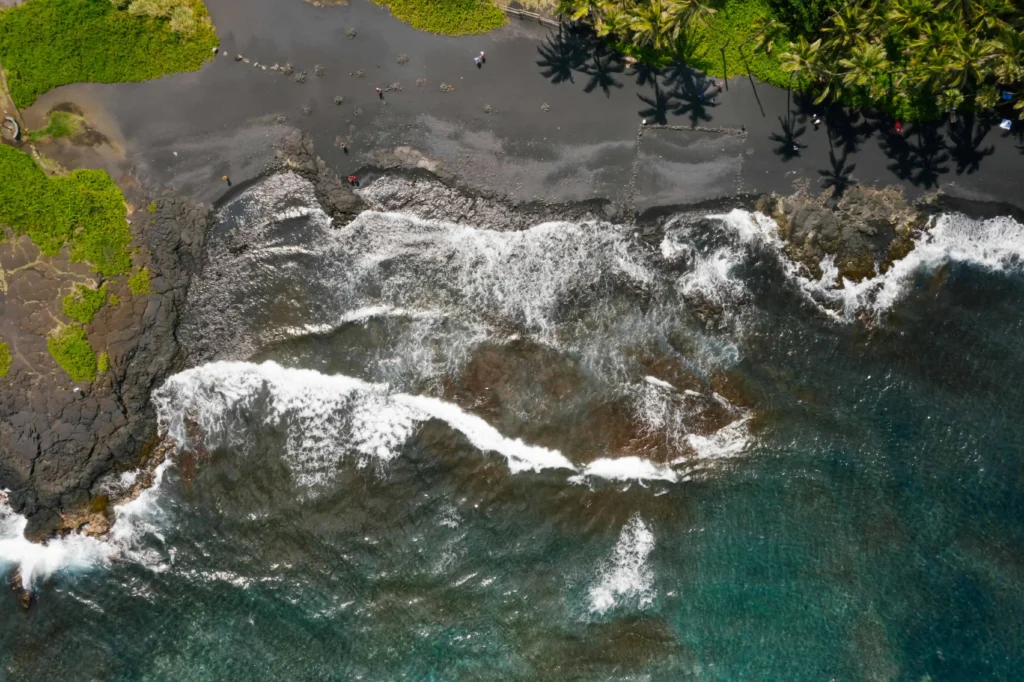 Aerial shot of a rugged black sand coastline with ocean waves crashing against the shore and palm trees nearby.