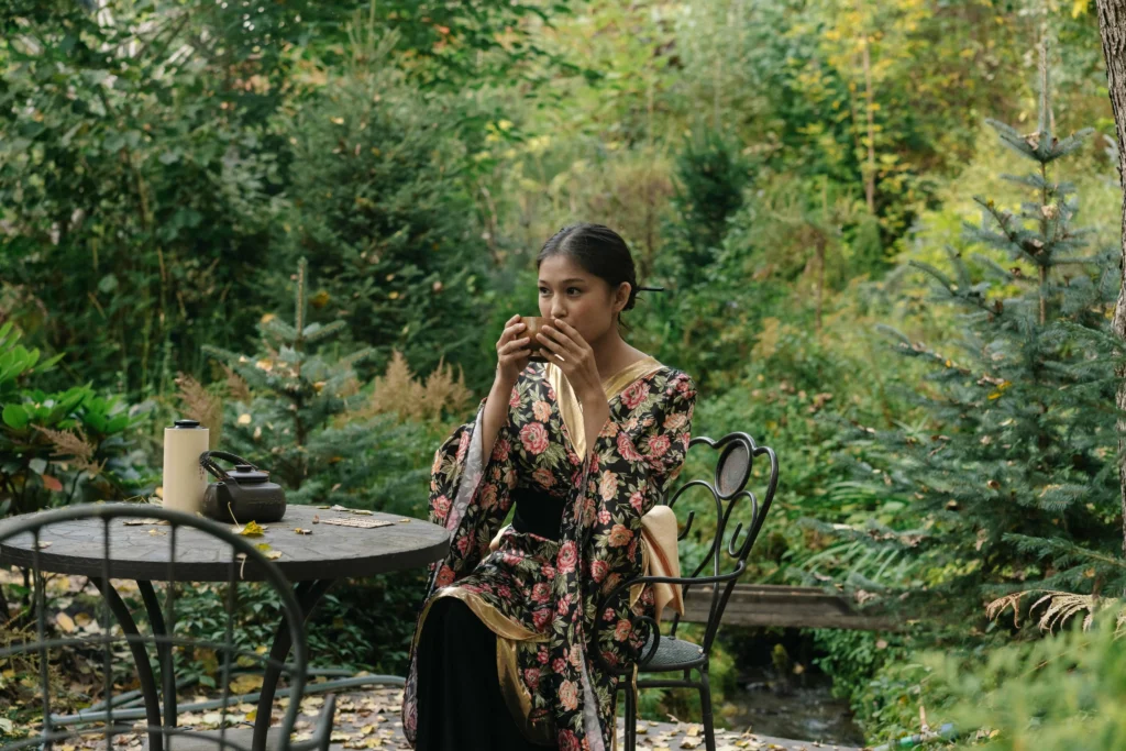 A woman in traditional floral attire sipping tea at a rustic outdoor table surrounded by vibrant greenery, enjoying a peaceful moment in nature
