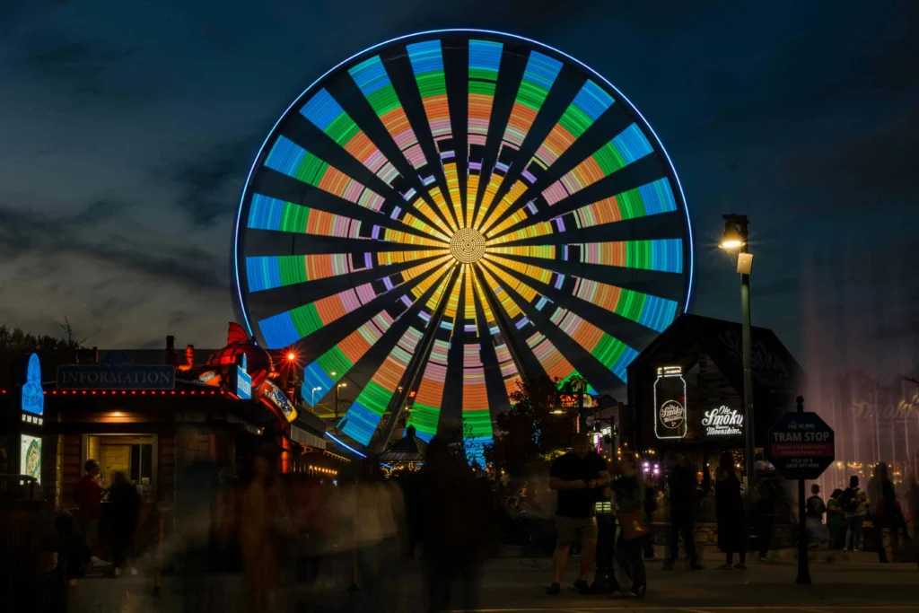 A vibrant, illuminated Ferris wheel at night, radiating colorful patterns against a dark sky, with lively amusement park activity and neon-lit shops in the foreground.