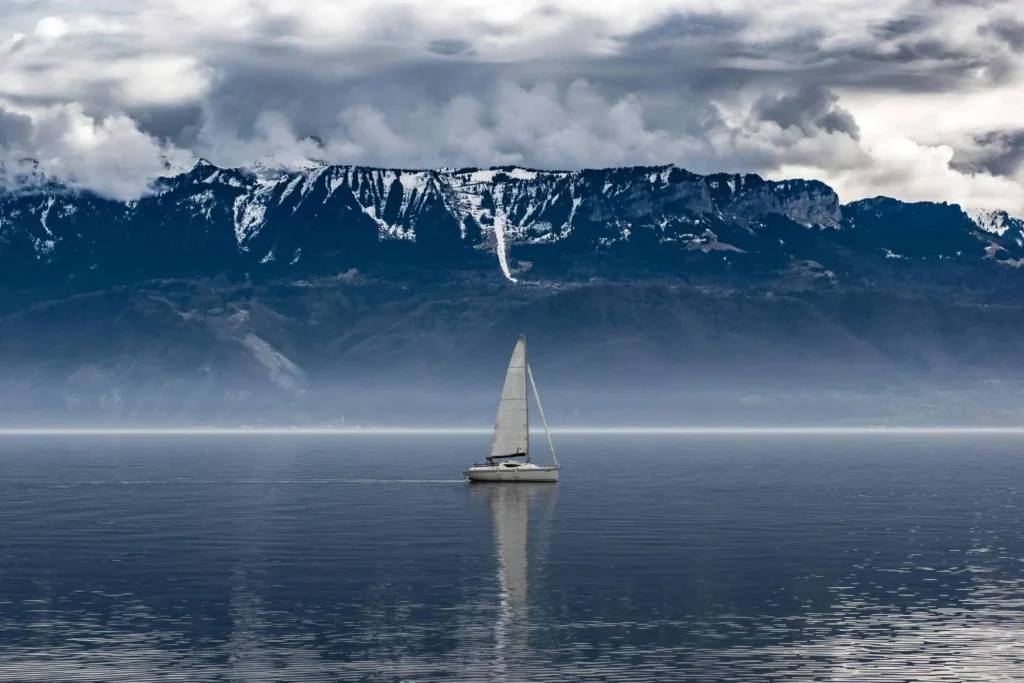 A serene sailboat gliding across the calm waters of Lake Geneva, Switzerland, under a dramatic cloudy sky. The majestic snow-capped Alps form a striking backdrop, creating a tranquil and picturesque alpine scene.