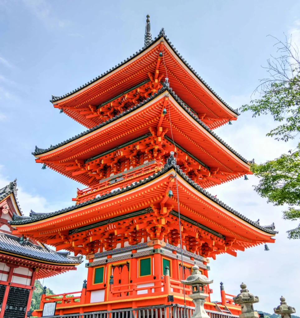 A vibrant orange three-tiered pagoda with intricate details, part of the Kiyomizu-dera Temple complex in Kyoto, Japan, set against a clear blue sky."