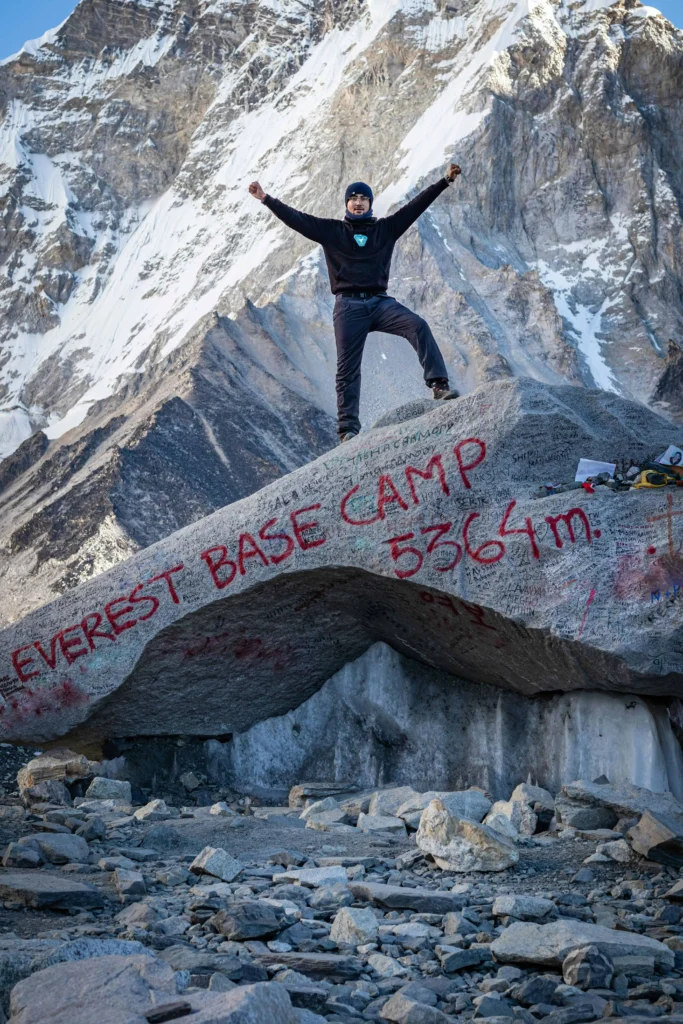 A climber triumphantly standing on a rock at Everest Base Camp, marked with "Everest Base Camp 5364m" in bold red letters, with the Himalayan mountains in the background.