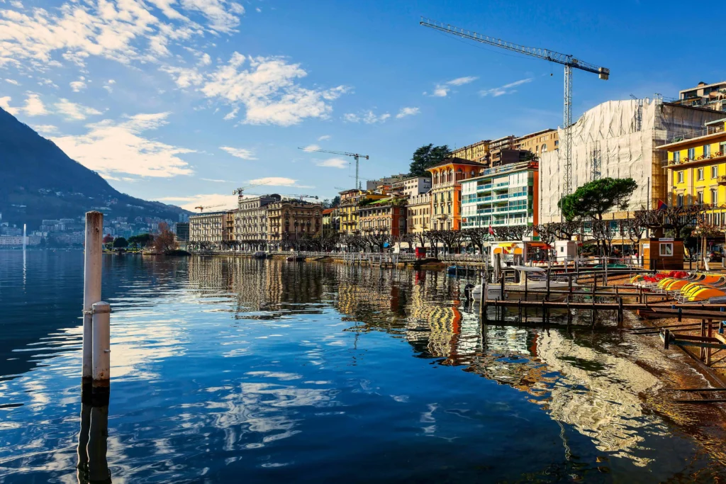 A vibrant lakeside view of Lugano, Switzerland, featuring colorful buildings along the waterfront reflecting in the calm blue waters of Lake Lugano. The scene is complemented by the silhouette of mountains in the background under a clear, sunny sky.