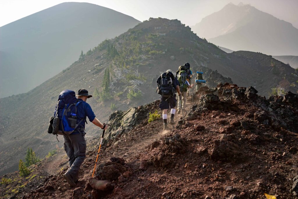 Hikers making their way up a steep, rocky mountain trail, with a large backpacker leading the group under the morning light."