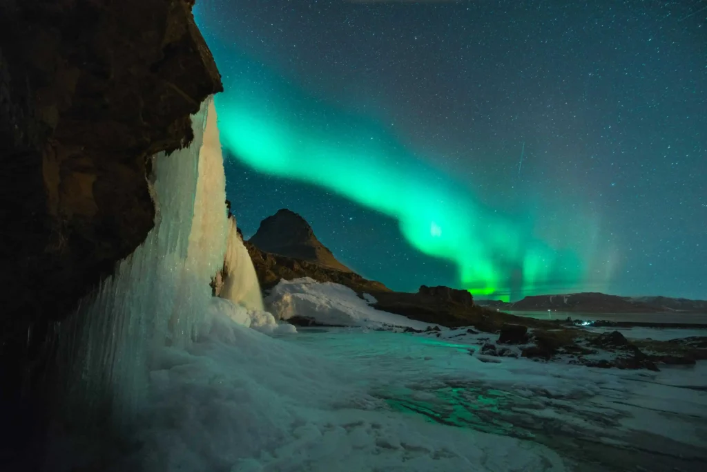 Iconic Kirkjufell mountain with vivid green northern lights reflecting on icy terrain in Iceland.