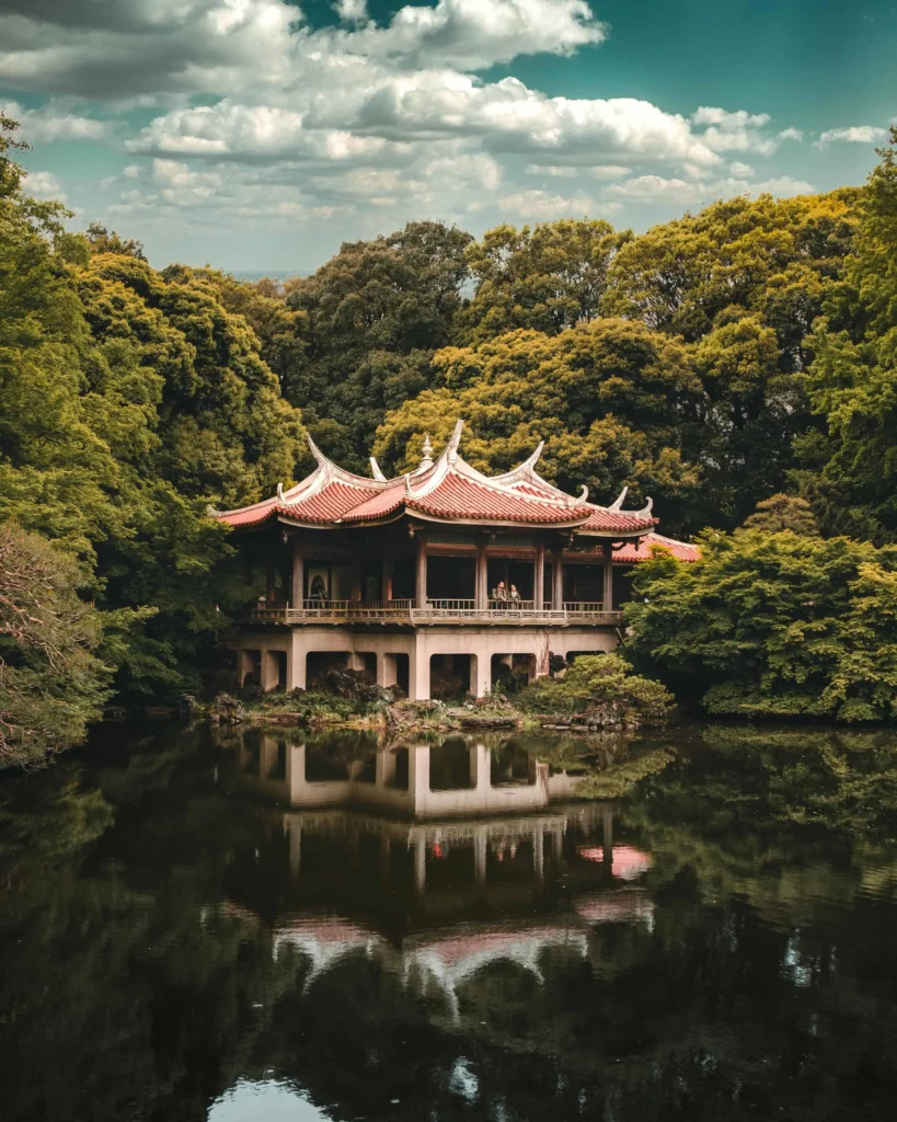 A traditional Japanese pavilion with intricate curved roofs, surrounded by lush green trees and reflected beautifully in a serene pond under a partly cloudy sky.