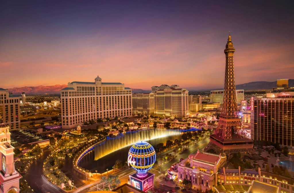 Panoramic view of the Las Vegas Strip at sunset, featuring the Eiffel Tower replica at Paris Las Vegas, the Bellagio fountains, and illuminated hotel buildings against a colorful sky