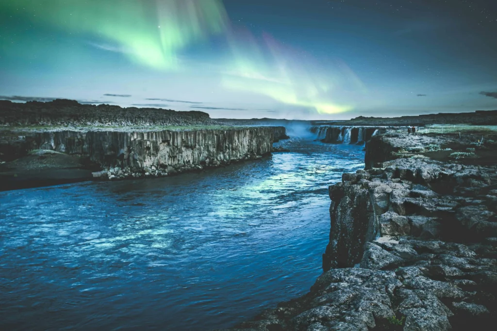 Northern Lights illuminating the sky over a serene river and rugged cliffs in Iceland, showcasing the country’s natural beauty and ideal conditions for aurora viewing