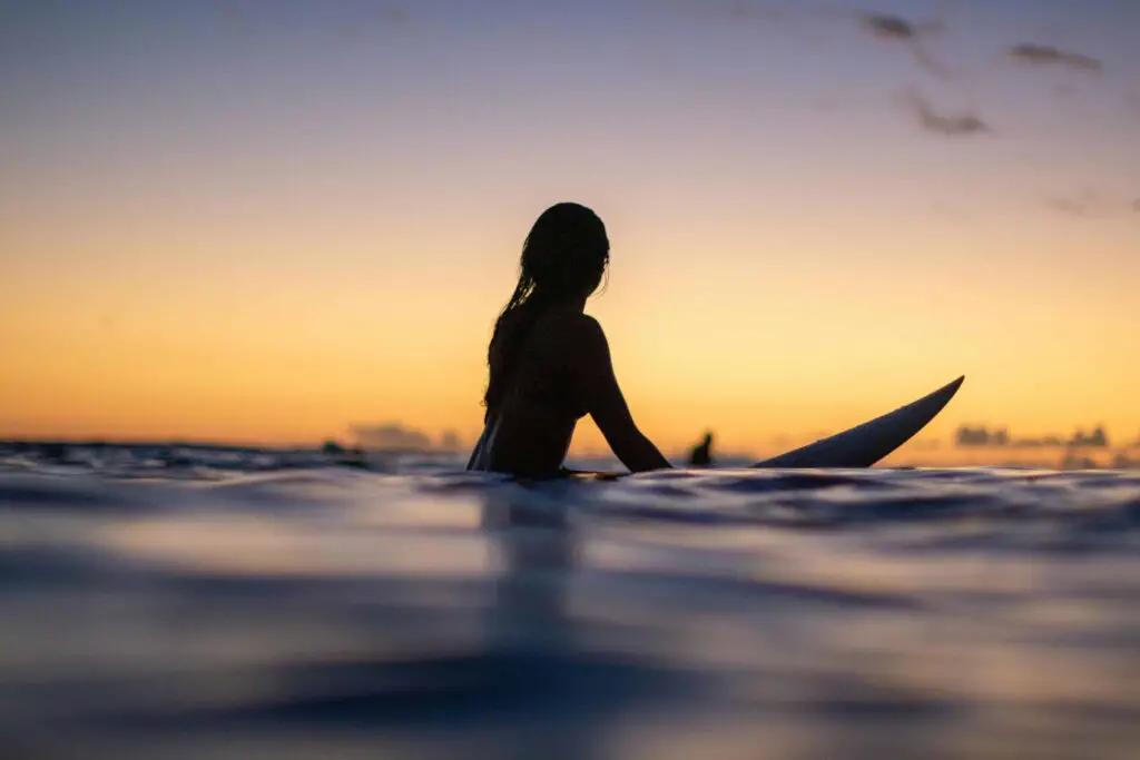 Silhouette of a surfer sitting on their board, gazing at the horizon during a serene sunset over the ocean.