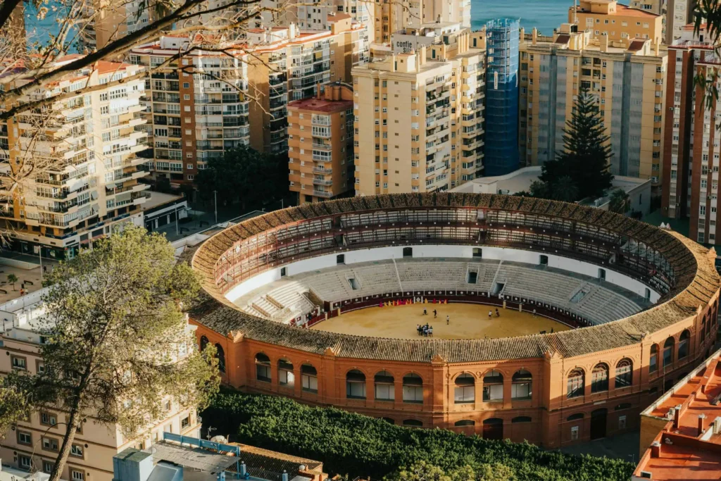 An aerial view of a historic bullring in Málaga, Spain, surrounded by modern apartment buildings and greenery, with a glimpse of the Mediterranean Sea in the background.