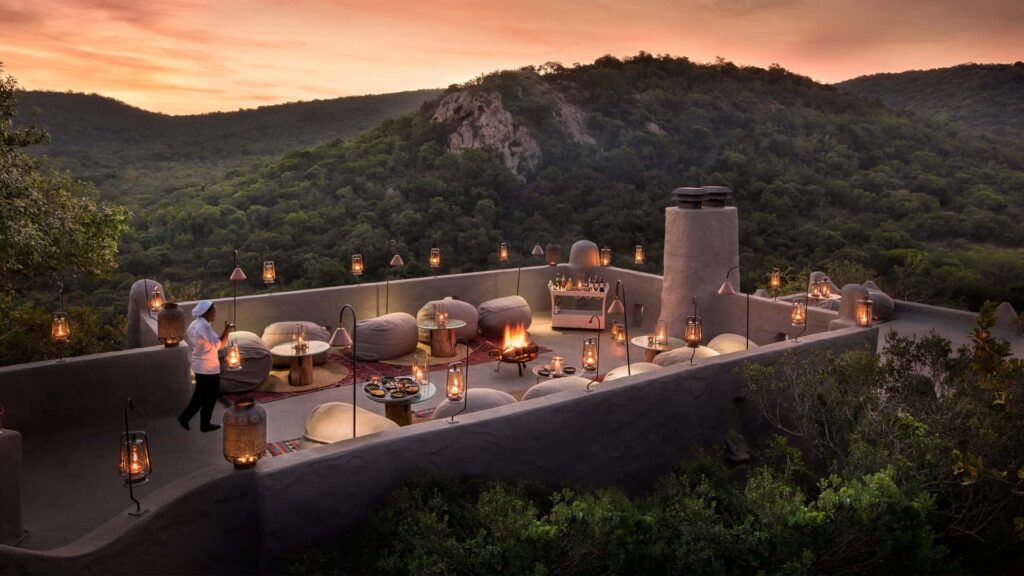 Romantic rooftop dining setup at Phinda Rock Lodge, featuring cozy seating, lanterns, and a warm fire pit surrounded by lush, green hills at sunset.