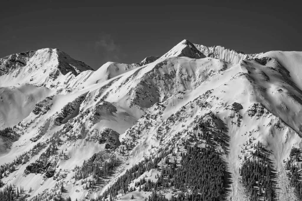 Snow-covered mountain peaks with pine trees scattered along the slopes under a clear sky, showcasing a serene winter landscape