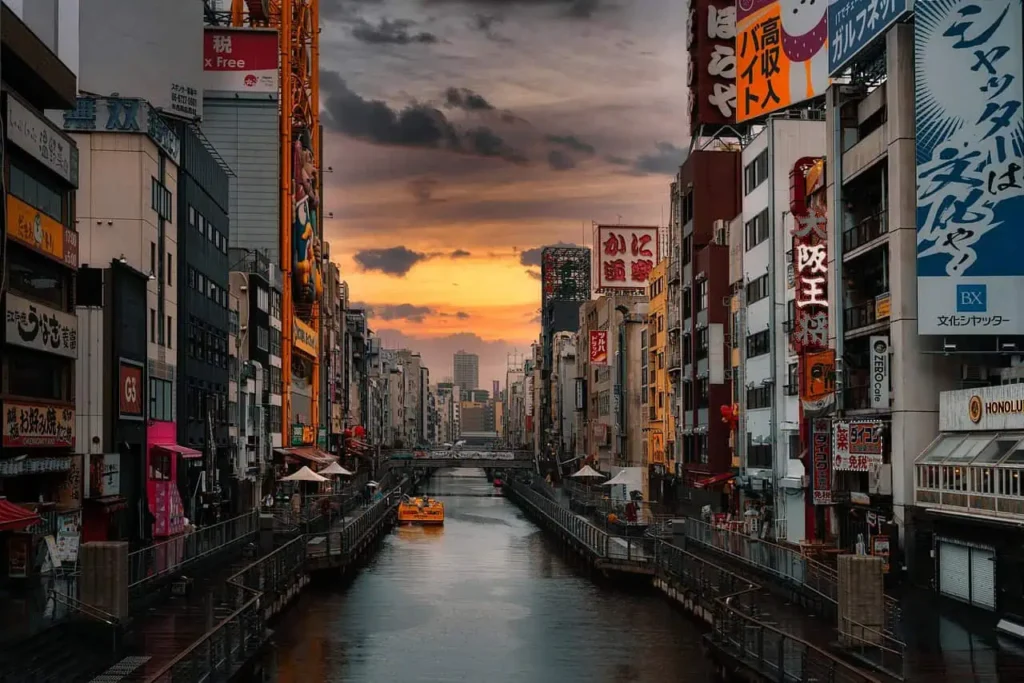 Vibrant Dotonbori district in Osaka at sunset, featuring a canal lined with illuminated billboards, restaurants, and a traditional sightseeing boat.