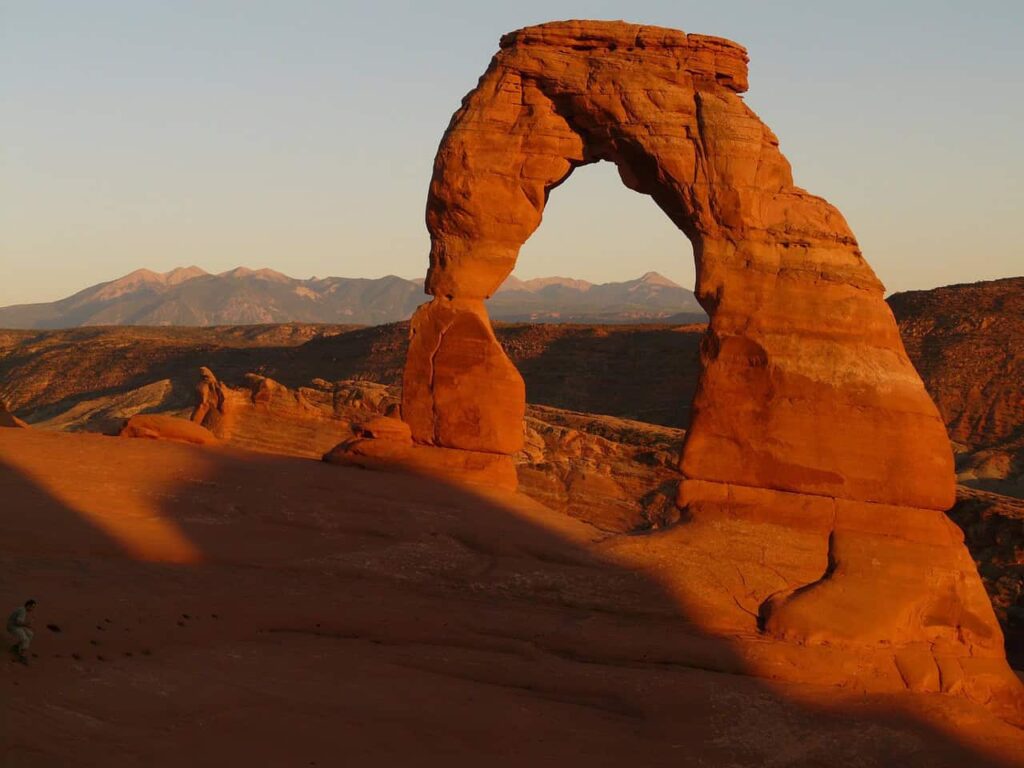 Delicate Arch at Arches National Park illuminated by the soft golden light of sunset, with the distant mountains in the background, highlighting the park’s unique sandstone formations.