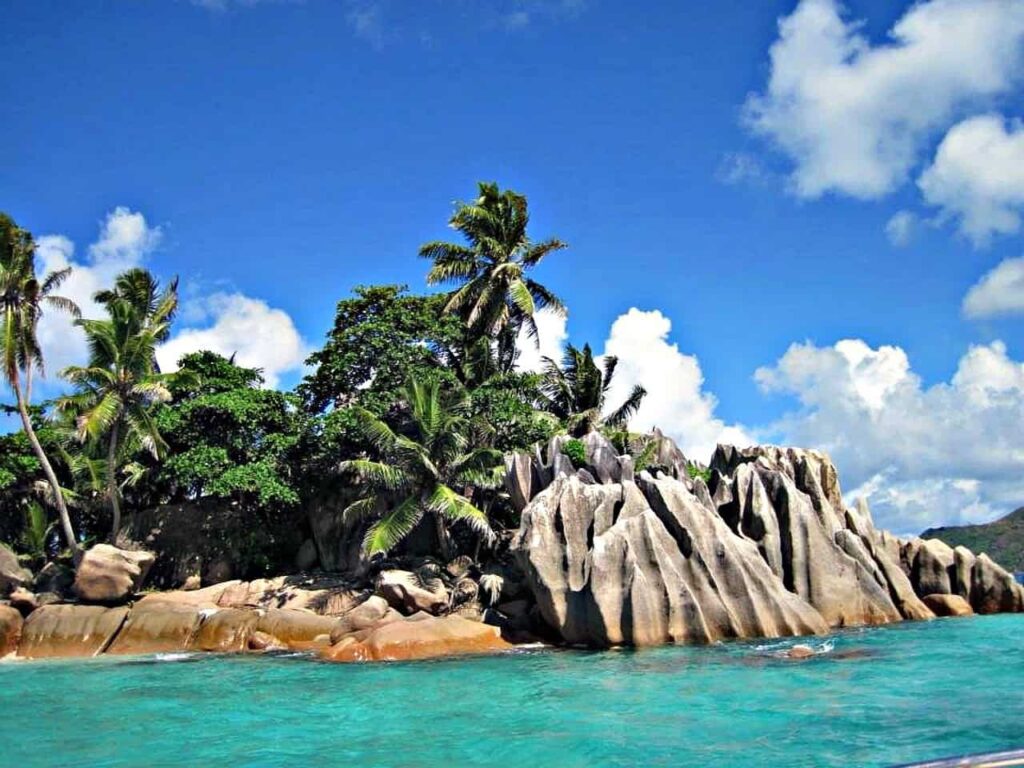 Scenic view of a stunning granite rock formation surrounded by lush green palm trees and crystal-clear turquoise waters under a bright blue sky in the Seychelles.