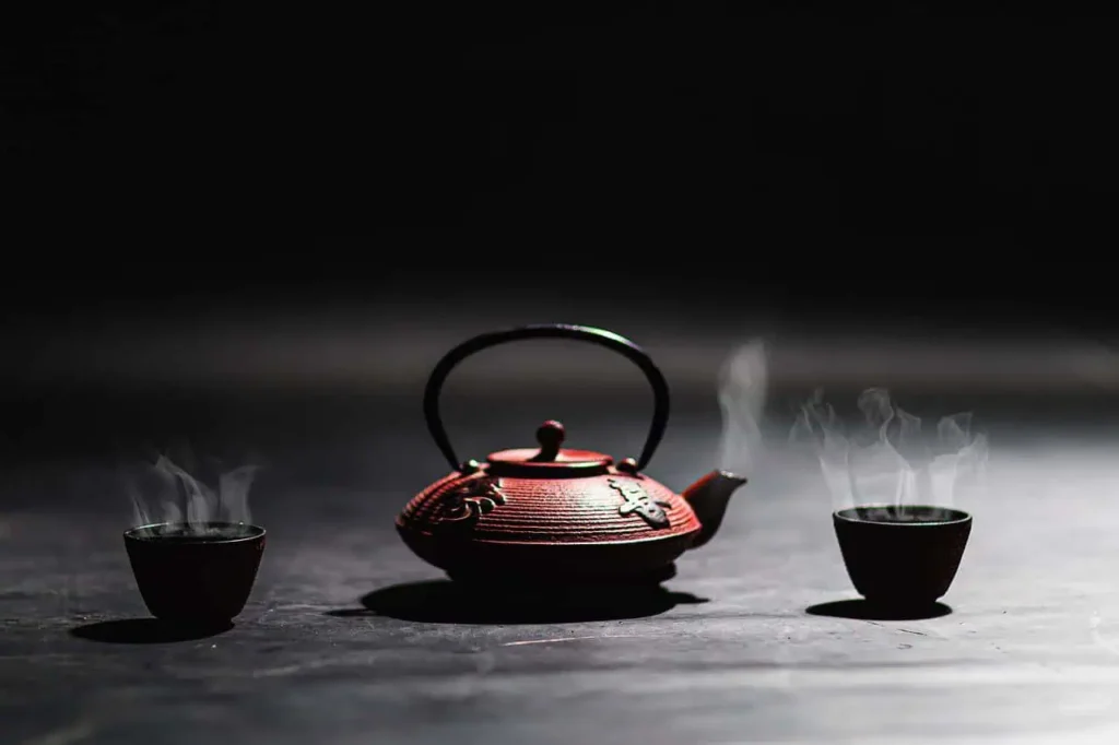 A steaming red Japanese teapot accompanied by two small teacups on a dark minimalist background, evoking warmth and tranquility.