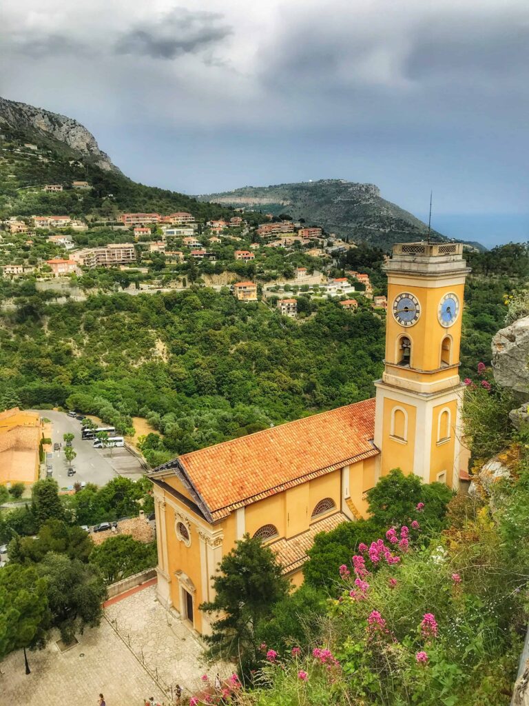 A picturesque view of Notre-Dame de l'Assomption Church in Eze, France, featuring its striking yellow façade and clock tower. The church is surrounded by lush greenery and vibrant pink flowers, with hills and the Mediterranean Sea visible in the distance under a cloudy sky.