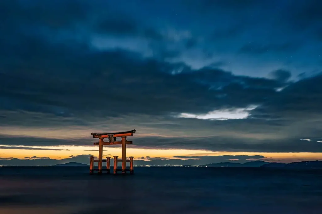 An iconic torii gate standing in the waters of Lake Biwa, Japan, silhouetted against a dramatic twilight sky with distant city lights on the horizon