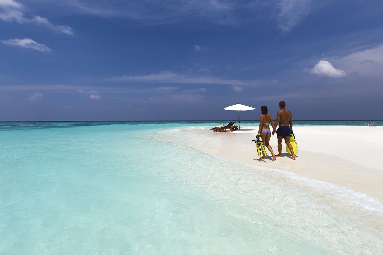 Couple walking hand in hand on a pristine white sandy beach, with clear turquoise waters and a beach umbrella in the background, symbolizing a romantic escape to one of the 10 best honeymoon lodges in Africa in 2025.