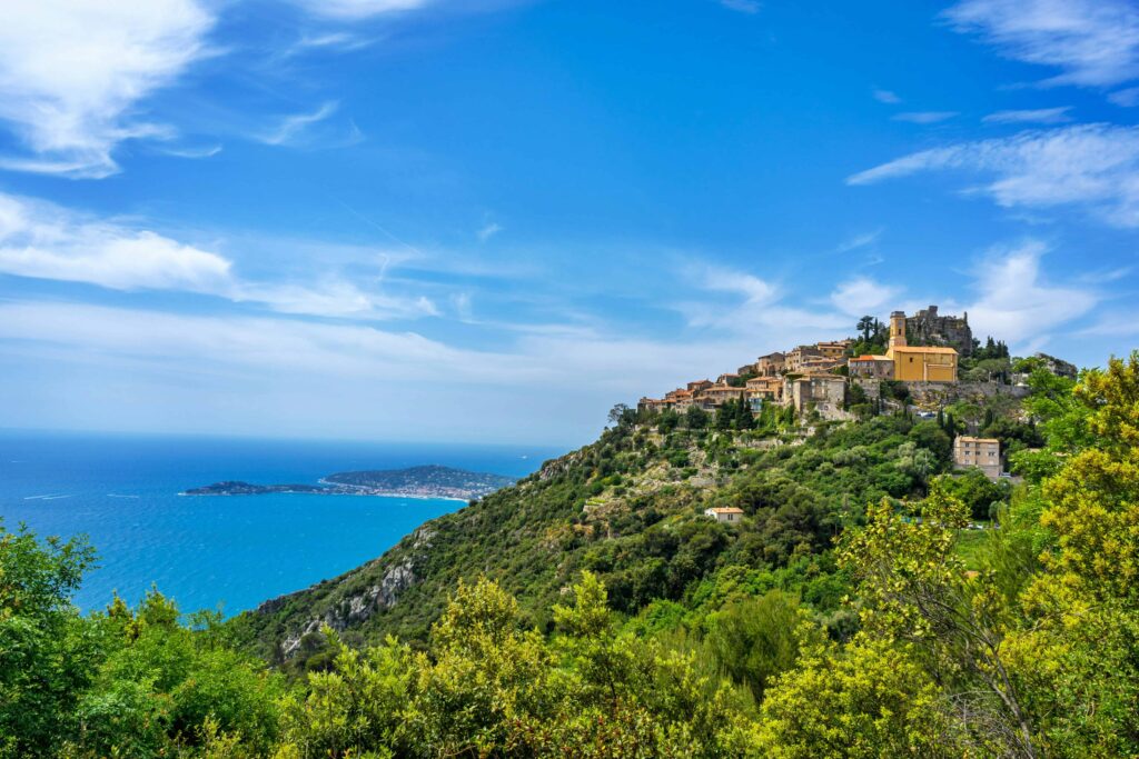 Stunning panoramic view of Eze, France, showcasing the picturesque medieval village perched on a hilltop surrounded by lush greenery, with the bright blue Mediterranean Sea stretching into the horizon under a clear, sunny sky