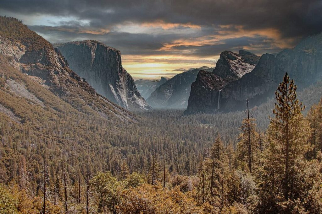 A dramatic view of Yosemite National Park's valley, with towering granite cliffs and a forested landscape under a cloudy, moody sky at sunset.