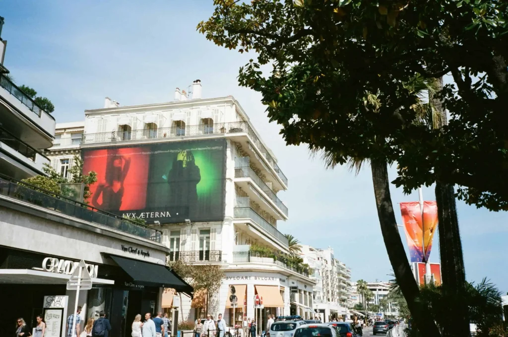 A lively street scene in Cannes, France, featuring luxury stores like Bulgari and Van Cleef & Arpels, with a prominent billboard displaying 'Lux Æterna' artwork. The street is lined with palm trees, bustling pedestrians, and cars under a clear blue sky, embodying the elegance and vibrancy of Cannes