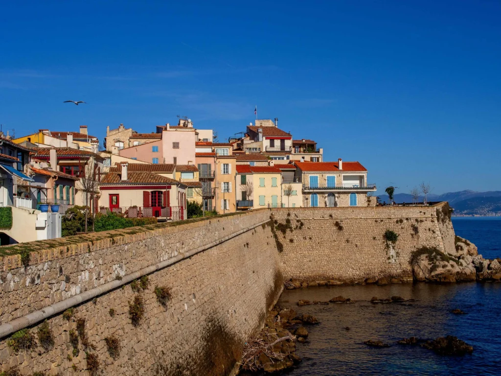 A charming coastal village with pastel-colored houses overlooking a stone wall and rocky shoreline, under a clear blue sky