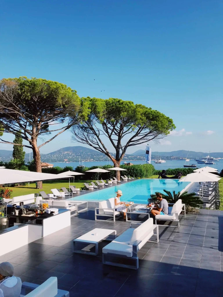 A luxurious poolside setting with modern white furniture, shaded by tall pine trees and overlooking a serene bay dotted with yachts in Antibes South of France