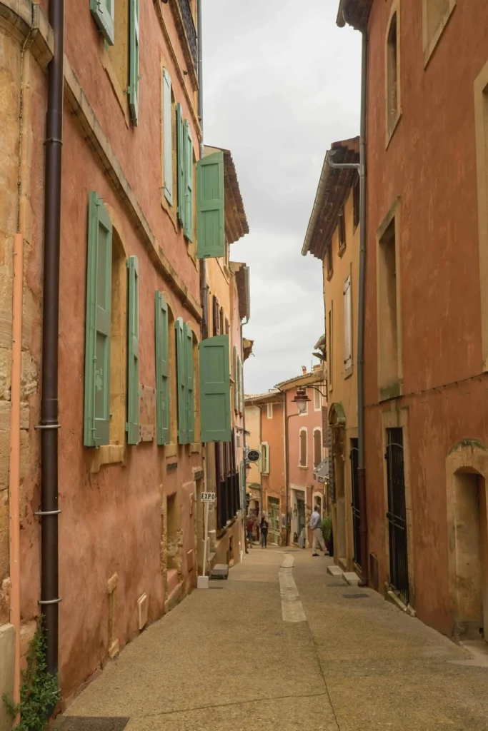 A narrow cobblestone street in Provence, flanked by pastel-colored buildings with vibrant green shutters and rustic charm.