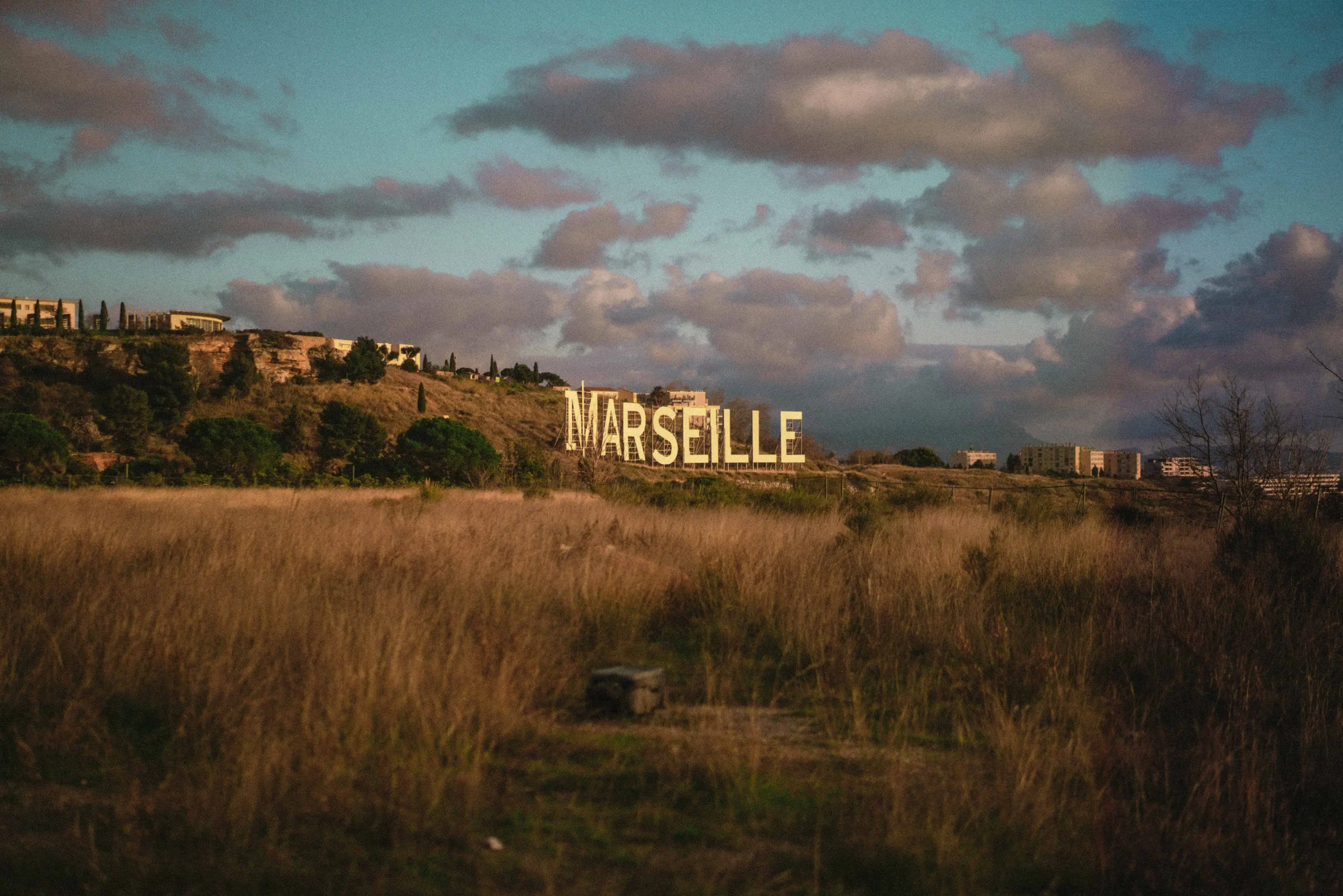 A large white "Marseille" sign on a hillside, reminiscent of the Hollywood sign, set against a backdrop of dramatic clouds and warm evening light.