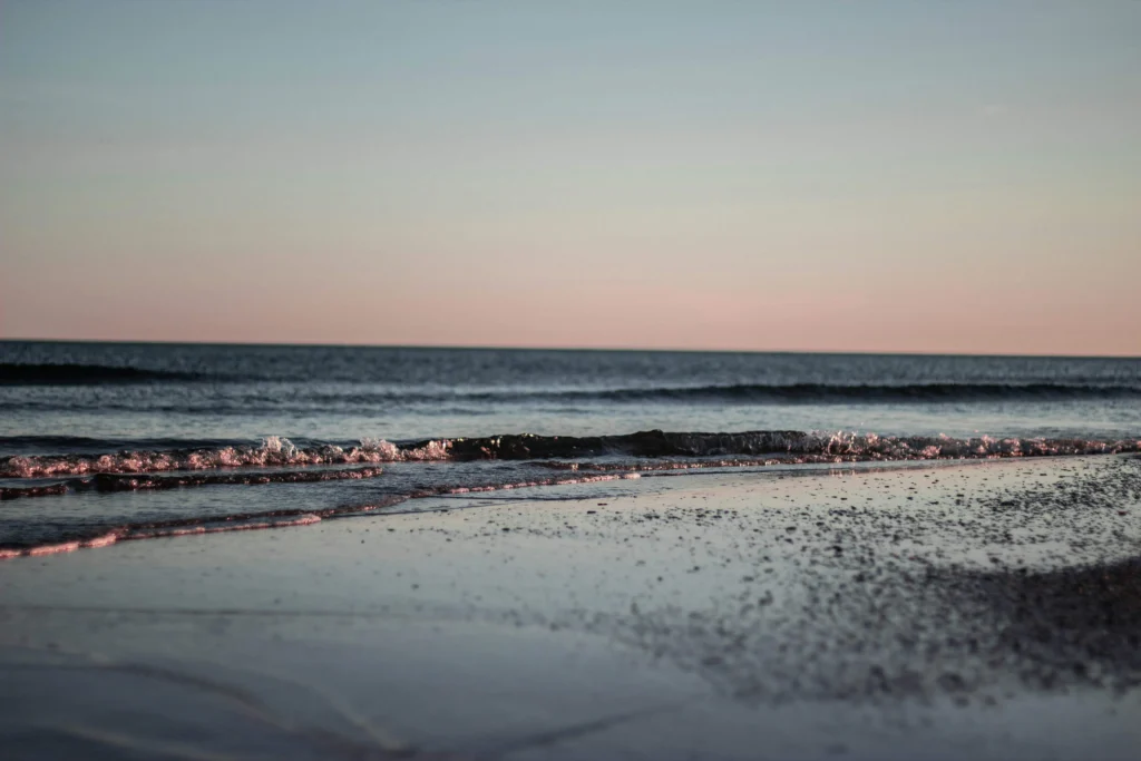 Gentle waves lapping at a serene sandy beach at sunset, with pastel skies reflecting on the water.