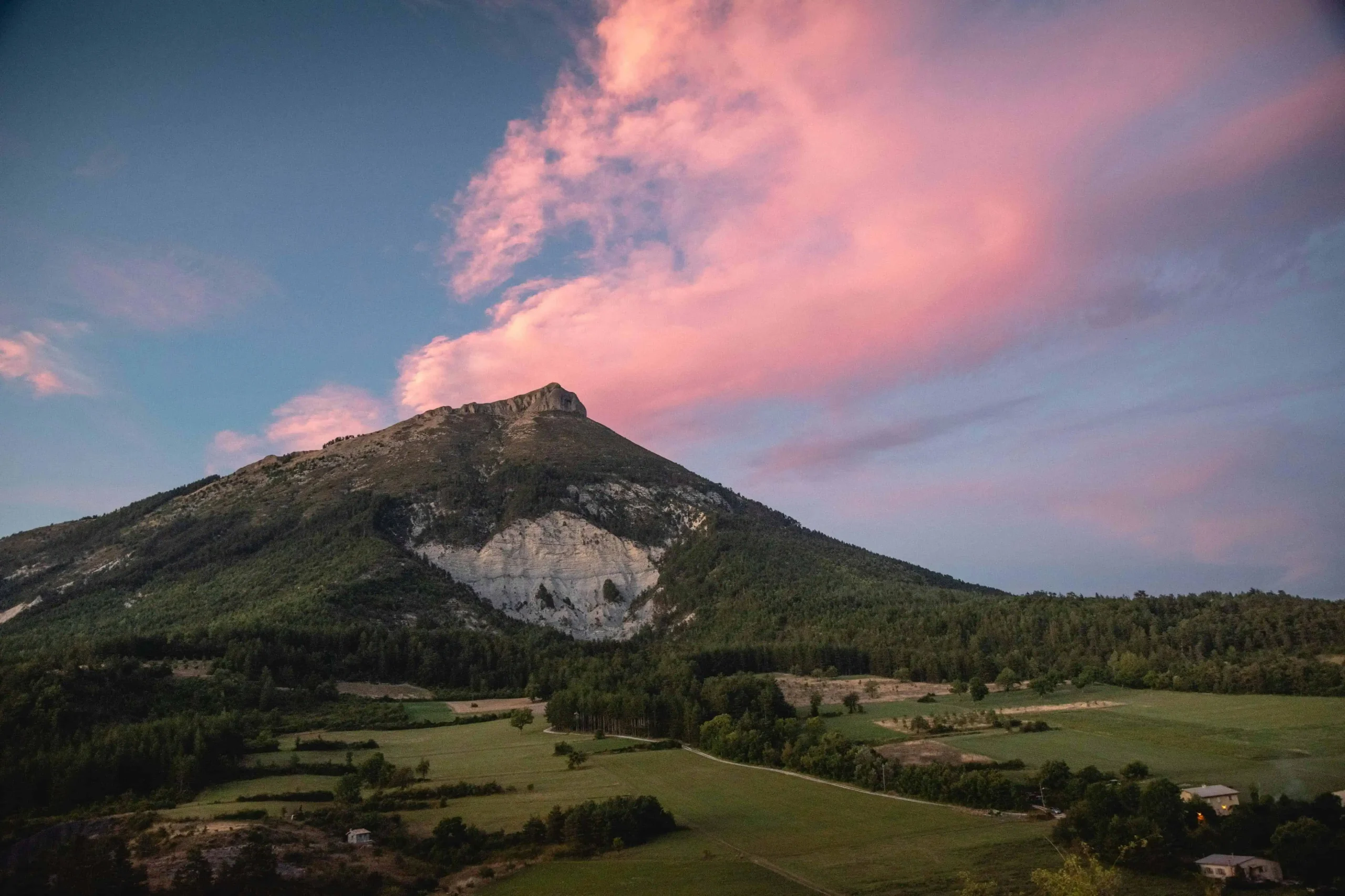 A dramatic mountain landscape in Languedoc, with pink clouds hovering above lush green fields at sunset.